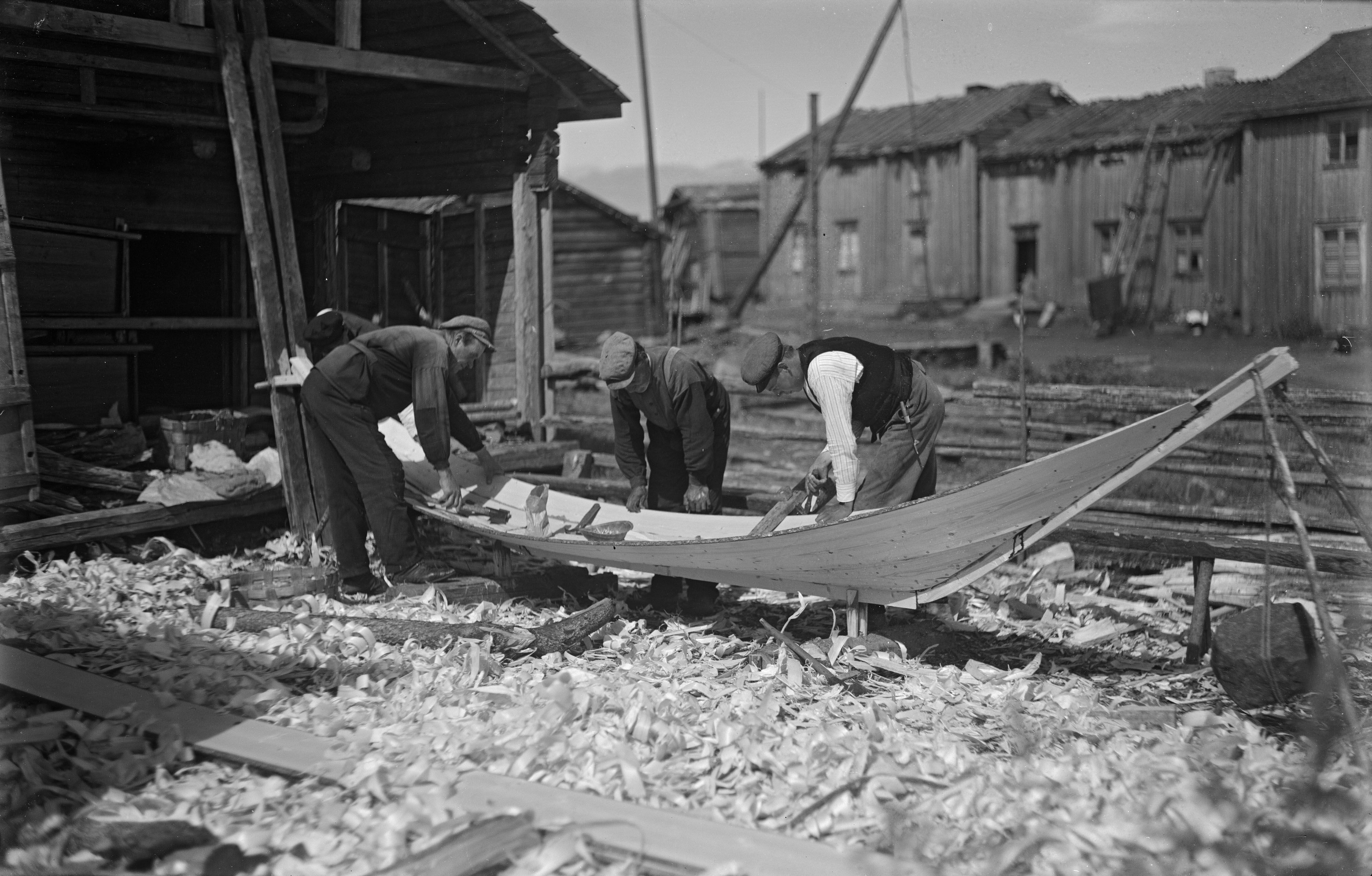 SLS 405a_30 Lillbåt byggs i Korsnäs. Fin utomhusbild med fyra båtbyggare i full fart. Massor av sågspån på marken och en intressant rad av hus i bakgrunden. Foto av Valter Forsblom 1930.